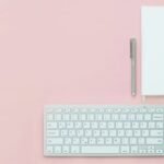 Flat lay of a minimalist workspace featuring a keyboard, notebook, and pen on a pink surface.